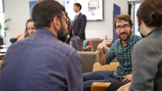 Paulo Savaget Paulo Savaget sitting with a group of people gesticulating and talking, wearing a green check shirt.