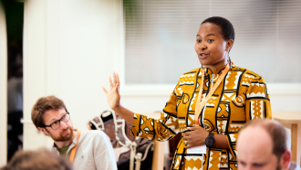 Anne Makena  Anne Makena wearing an orange patterned dress presenting to a crowd at the Skoll Community Convening.
