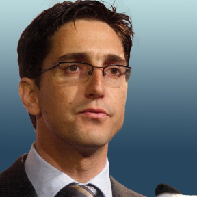 François Bonnici   François Bonnici  in front of a blue background wearing a beige suit and tie.